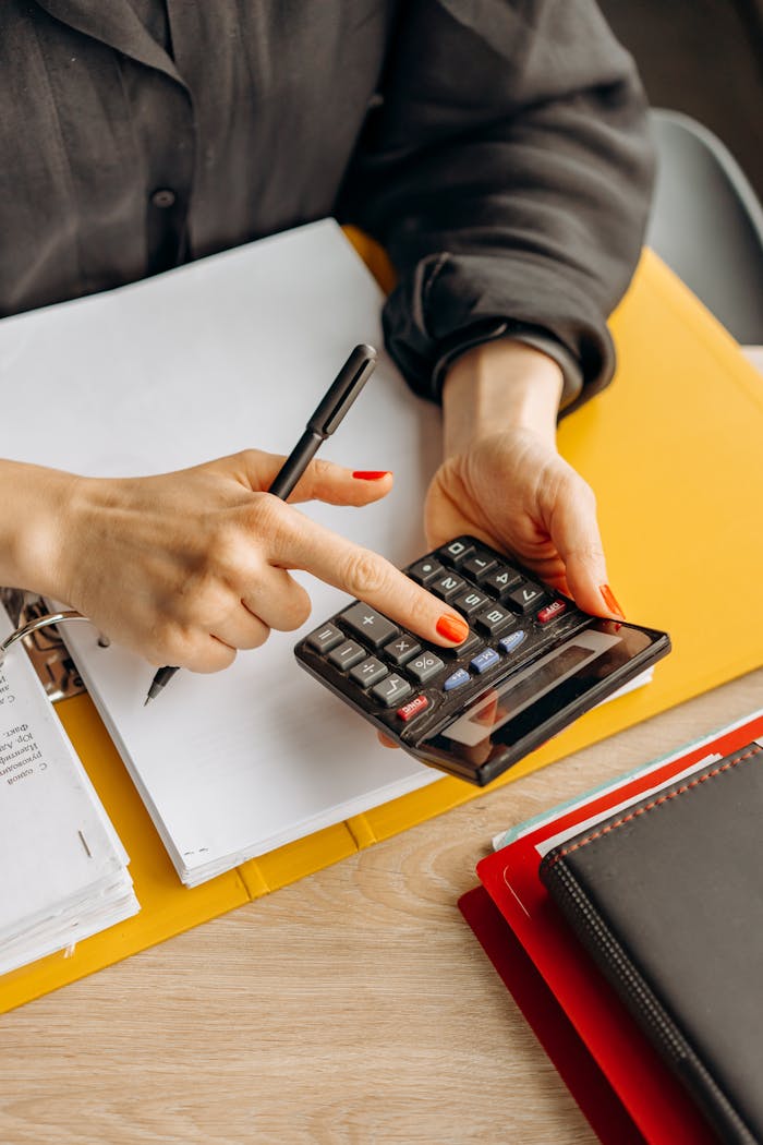 Close-up of a woman using a calculator at her wooden workspace with documents and notepads.
